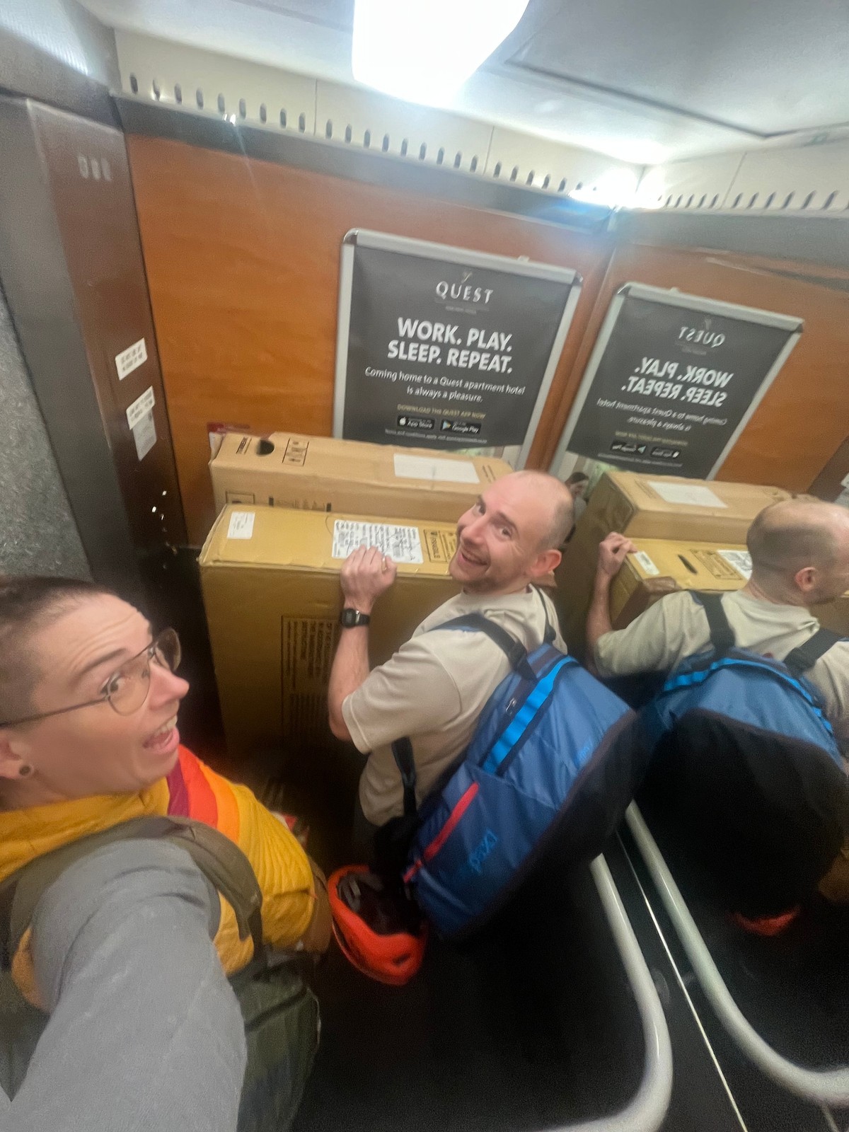 Two individuals are crammed into a small elevator, each holding onto a large cardboard box while wearing backpacks. They both smile at the camera, surrounded by the elevator’s wooden paneling and a promotional poster for a hotel.